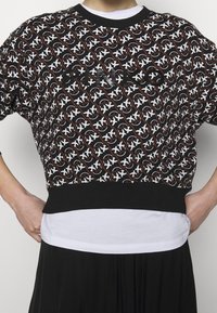 Czarnobiały sweter w wzory z okrągłym dekoltem, krótkimi rękawami i elastycznym pasem. Pod spodem zakrzywiona biała koszula.