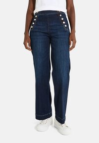 Jean en denim bleu taille haute avec des jambes larges, présentant des accents de boutons sur le côté et une texture légèrement usée. Associé à des baskets blanches.