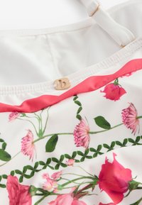 Tessuto bianco con stampa floreale rosa e foglie verdi, con bordatura rossa e piccolo dettaglio logo dorato vicino allo scollo su una gruccia bianca.