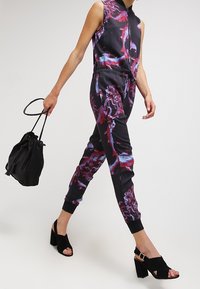 Svart jumpsuit med flerfärgade abstrakta mönster, med dragkedja fram, dragsko i midjan och ribbade muddarf. Kombinerad med svarta klackskor.