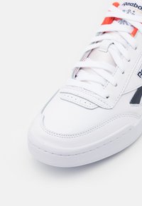 Gros plan sur une sneaker Reebok Classic blanche avec des lacets blancs, une doublure de languette rouge et des détails de logo noirs sur un fond blanc.