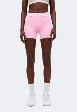 Personne athlétique portant un short Nike Pro rose, un soutien-gorge de sport blanc, des chaussettes blanches montantes et des baskets Nike blanches, debout, bras détendus.