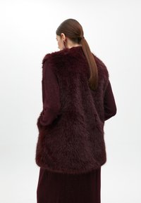 Burgundské kožichové vesta s dlouhým, texturovaným designem, nošená přes odpovídající šaty. Hladká tkanina pod tím, s jemným leskem.
