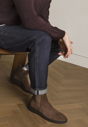 Bottes de cheville en daim marron avec des panneaux élastiques sur les côtés, portées avec un jean en denim foncé retroussé. Semelle lisse et plate avec un design simple. Fond en bois.