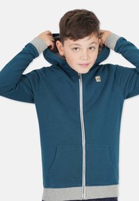 Blå zip-hoodie med grå insida och muddar, som har en framficka och en liten logotyp på vänster bröst.