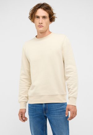 Jasnobeżowa bluza z długim rękawem o okrągłym dekolcie, z prążkowanymi ściągaczami na rękawach i u dołu, z małym logo na piersi. Noszona z niebieskimi dżinsami.