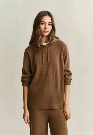 Jonge vrouw met lang bruin haar, die een bruine gebreide hoodie en bijpassende broek draagt, staand tegen een effen beige achtergrond.