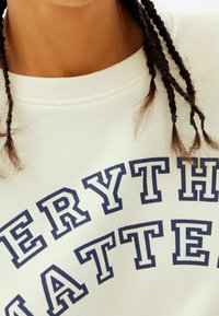 Vit sweatshirt med djärv marinblå text som står "ALLT ÄR VIKTIGT." Ribbad halsringning och texturdetaljer synliga.
