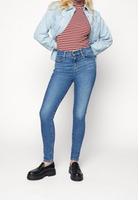 Jeans skinny de mezclilla azul claro con cintura alta, combinados con un suéter de cuello alto a rayas rojas y blancas y una camisa de botones azul claro. Zapatos negros.