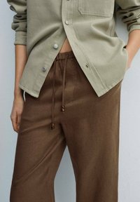 Pantalon en lin marron avec une taille à cordon, orné d'accents en perles de bois. Associé à une chemise boutonnée vert clair.