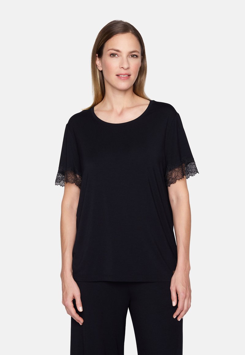 Frau mit glattem, schulterlangem Haar, die ein schwarzes Kurzarmoberteil mit Spitzenbesatz an den Ärmeln trägt, vor einem schlichten weißen Hintergrund.