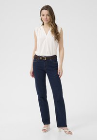 Blouse blanche à manches courtes avec des bords volantés, associée à un jean droit bleu marine et une ceinture brune. Le modèle porte des talons blancs à lanières.