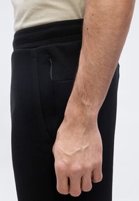 Κοντινό πλάνο του δεξιού χεριού ενός ατόμου που ακουμπάει δίπλα σε μαύρο παντελόνι με φερμουάρ στην πλαϊνή τσέπη και μέρος μιας μπεζ πουκαμίσας φαίνεται από πάνω.