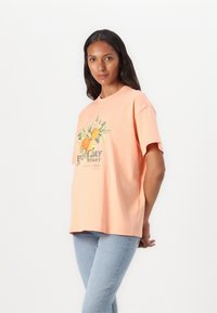 Camiseta de algodón color durazno con un gráfico floral que incluye rodajas de naranja y texto, mangas cortas y un ajuste holgado.