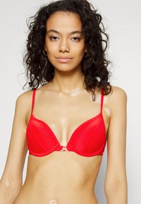 Sutiã vermelho com aro, textura canelada, alças ajustáveis e um logotipo em tom dourado no centro.