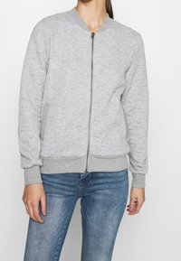 Grijze bomberjas met ritssluiting aan de voorkant, een textuurpatroon, elastische manchetten en tailleband. Draagt over een blauwe spijkerbroek, met zijzakken.