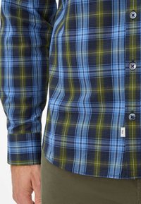 Kurzarm-Hemd aus Karostoff in Blau, Grün und Marineblau mit Knopfverschluss, das ein kariertes Muster und eine dezente Etikettenakzentuierung aufweist. Strukturierter Stoff.