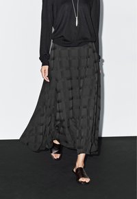 Zwarte lange rok met een gestructureerd geruit patroon, gecombineerd met een losse zwarte top en donkere slip-on sandalen. Soepele stof met een vloeiend ontwerp.