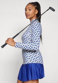 Golf dress in blue and white patterned fabric, featuring a high collar and a pleated blue skirt. Model holds a golf club over her shoulder.