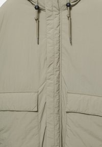 Svetlo béžová bunda s kapucňou, nastaviteľné šnúrky s čiernymi korálkami, vrecká na klopu a centrálny zips skrytý pod tkaninovým prekrytím.