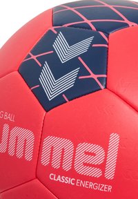 Röd och marinblå fotboll med hexagonala paneler, som har vita randiga detaljer och en texturerad yta. Varumärket säger "mel CLASSIC ENERGIZER."