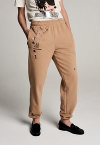 Beige Sweatpants mit schmalem Schnitt, die Farbspritzer und ein schwarzes Logo aufweisen. Hergestellt aus einem weichen, strukturierten Stoff mit einem elastischen Bund.