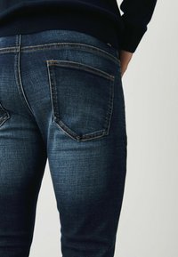 Jeans din denim de un albastru închis, cu o textură netedă, având două buzunare la spate și cusături portocalii în contrast de-a lungul cusăturilor.