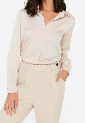 Kobieta nosząca jasnobeżową, zapinaną na guziki koszulę z długim rękawem, wpuszczoną w beżowe spodnie z wysokim stanem z czarnym guzikiem.