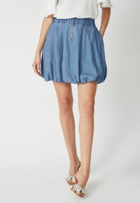 Denim ballon szoknya gumis derékkal, világos kék színben, középen cipzár részlettel, átlátszó sarkú szandálokkal párosítva.