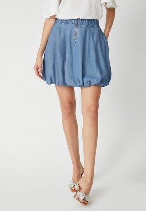 Gonna a palloncino in denim con elastico in vita, colore azzurro chiaro, e dettaglio della cerniera al centro, abbinata a sandali con tacco trasparenti.