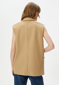Blazer beige senza maniche con colletto strutturato, texture liscia e cucitura centrale sulla schiena. Abbinato a jeans blu scuro.