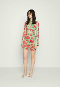 Mulher com cabelo escuro e comprido, a usar óculos com lente amarela, uma blusa de manga longa verde com padrão floral e recortes, saia a condizer e sandálias brancas.