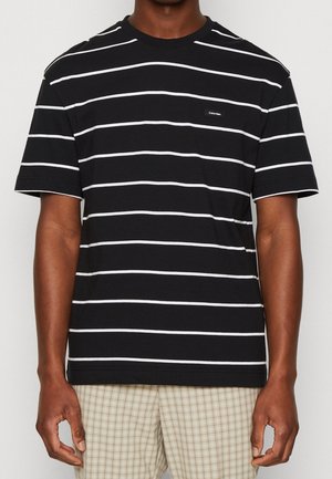 Mand iført en sort T-shirt med korte ærmer, hvide vandrette striber og et lille Calvin Klein-logo på brystet.