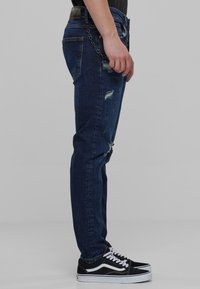 Vaqueros de mezclilla azul oscuro con un ajuste slim, que presentan detalles desgastados y un acento de cadena. Combinados con zapatillas negras y blancas.