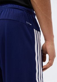 Gros plan sur une personne portant un short de sport bleu marine avec des rayures blanches verticales et le logo Climacool près de la taille.