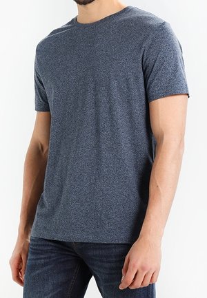 Mann trägt ein einfaches dunkelgraues T-Shirt mit kurzen Ärmeln und dunkelblaue Jeans, dargestellt von Hals bis Hüfte vor weißem Hintergrund.