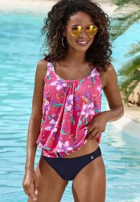 Roze tankini-top met groene en witte bloemen, gecombineerd met zwarte bikini-broekjes. Model met spiegende zonnebril; achtergrond bij het zwembad.