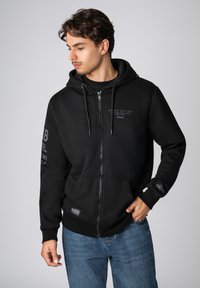 Zwarte hoodie met ritssluiting van zacht materiaal, voorzien van een kangoeroezak, trekkoorden en logo-accenten op de borst en mouw.