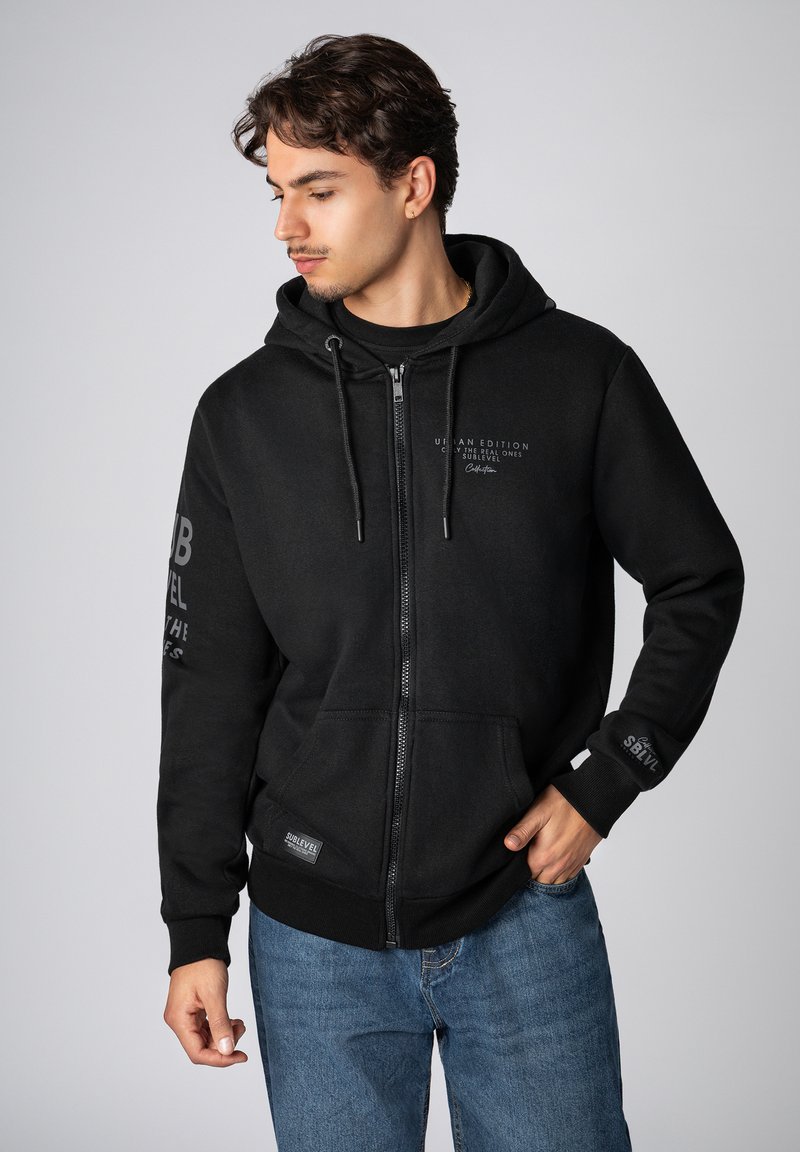 Zwarte hoodie met ritssluiting van zacht materiaal, voorzien van een kangoeroezak, trekkoorden en logo-accenten op de borst en mouw.