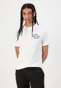 White short-sleeve t-shirt featuring a navy graphic text logo on the left chest, paired with black trousers. Smooth cotton material.