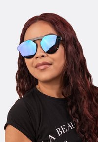 Black oversized sunglasses with blue mirrored lenses, featuring a geometric design and gold accents. Worn by a person with wavy hair.