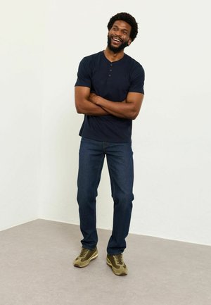 Tricou Henley cu mâneci scurte, culoare bleumarin, cu trei nasturi, asortat cu blugi denim albastru închis și adidași verde-măsliniu. Fundal simplu.