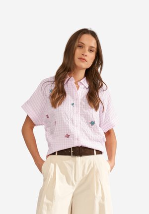 Naine, kes kannab roosat ruudulist lühikeste varrukatega särki väikeste tikanditega ja kreemikas volditud pükse tumeda vööga.