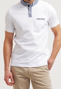 Homme portant un polo blanc à manches courtes avec un col gris et un détail de poche, associé à un pantalon beige.
