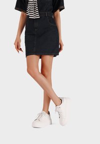 Donna che indossa una mini gonna in denim scuro, una sciarpa a righe bianche e nere, e delle sneaker bianche con la piattaforma, in piedi con le gambe incrociate.