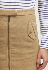 Jupe en laine beige avec une fermeture éclair et une poche décorative à boutons. Présente une texture lisse et une coupe ajustée. Chemise rayée visible au-dessus.