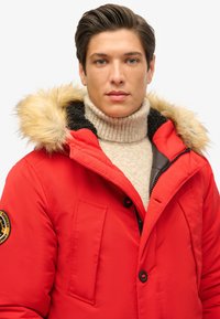 Czerwona kurtka z kapturem z sztucznego futra, czarną podszewką i dwiema przednimi kieszeniami. Noszona na beżowym swetrze z wysokim kołnierzem, z teksturowanym splotem.