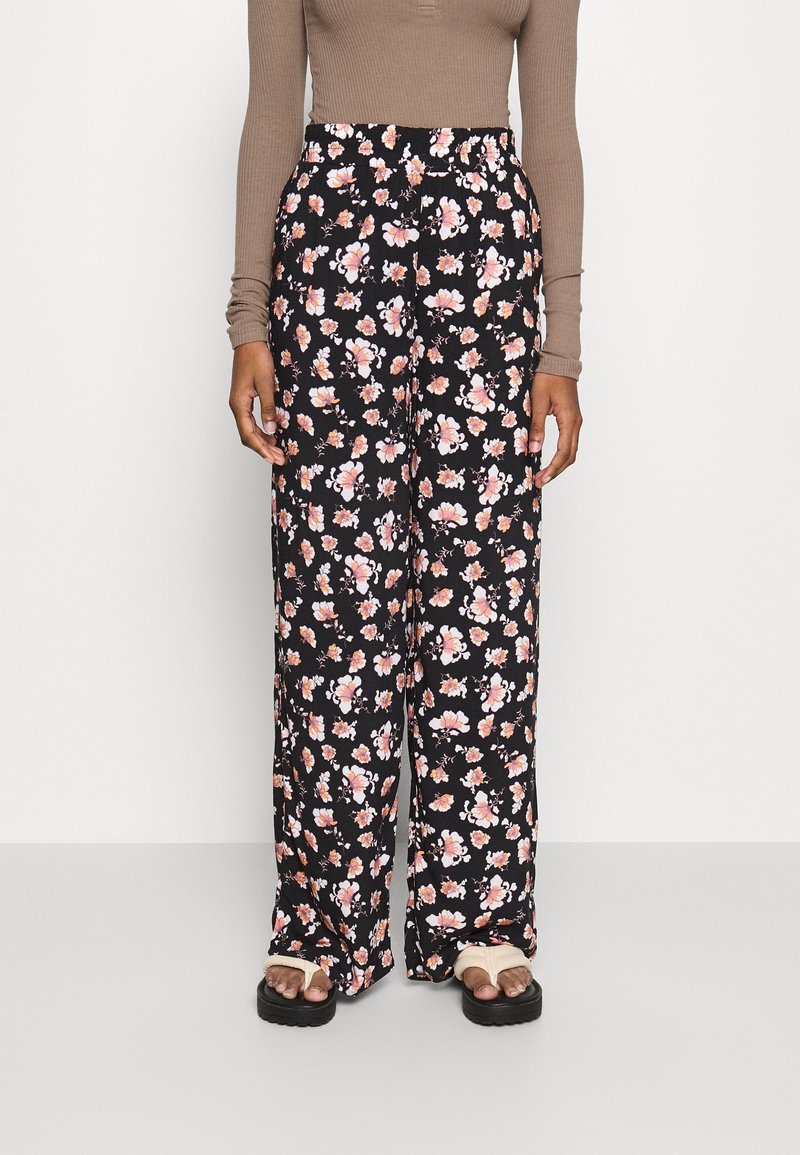 Personne portant un pantalon noir à jambes larges avec un imprimé floral rose et blanc, associé à un haut beige à manches longues et des sandales à plateforme beiges.