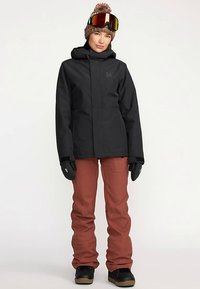 Chaqueta impermeable negra con capucha, combinada con pantalones de esquí color óxido. El modelo lleva un gorro de punto y gafas de esquí, con guantes en las manos.