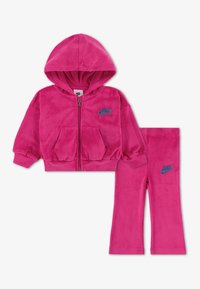Rozi velur Nike trenirka koja se sastoji od dukserice sa patentnim zatvaračem i odgovarajućih hlača, oboje s plavim logotipom i prednjim džepovima na dukserici.